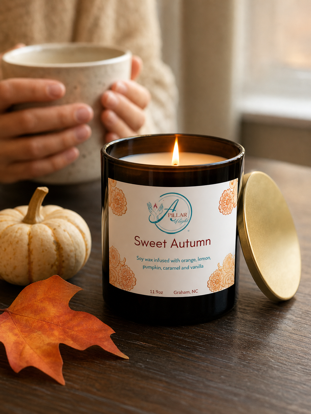 A lit amber glass candle jar labeled 'Sweet Autumn' by A Pillar of Light Candle Co., set on a wooden table with a gold lid beside it, a small pumpkin, an orange leaf, and a person holding a mug in the background.
