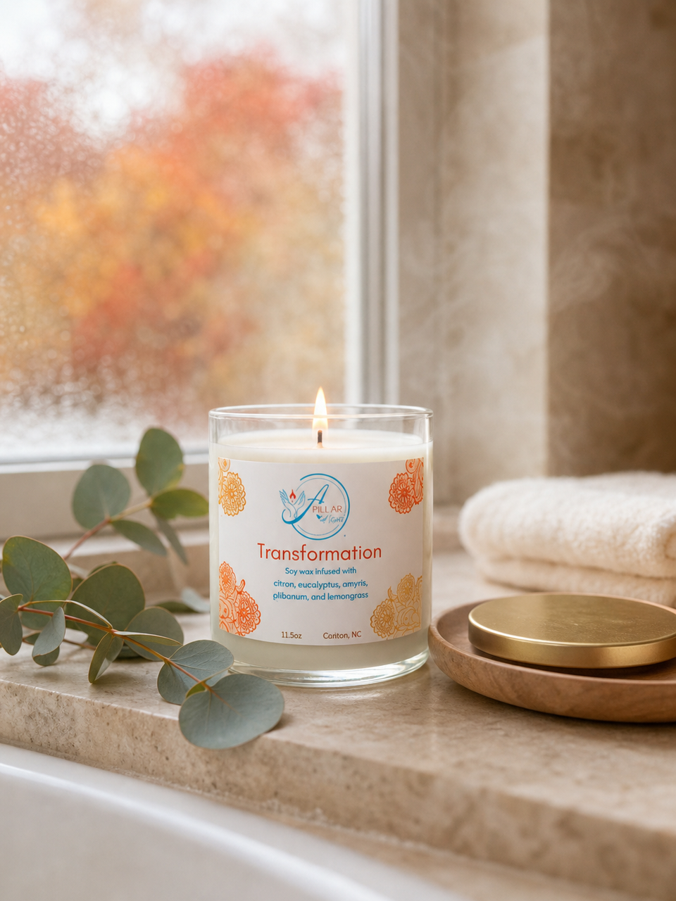A lit glass jar candle labeled 'Transformation' sits on a bathroom windowsill with a towel, gold lid, and eucalyptus leaves nearby. The candle is shown in a real interior setting, so this is Lifestyle rather than Product/Packaging.