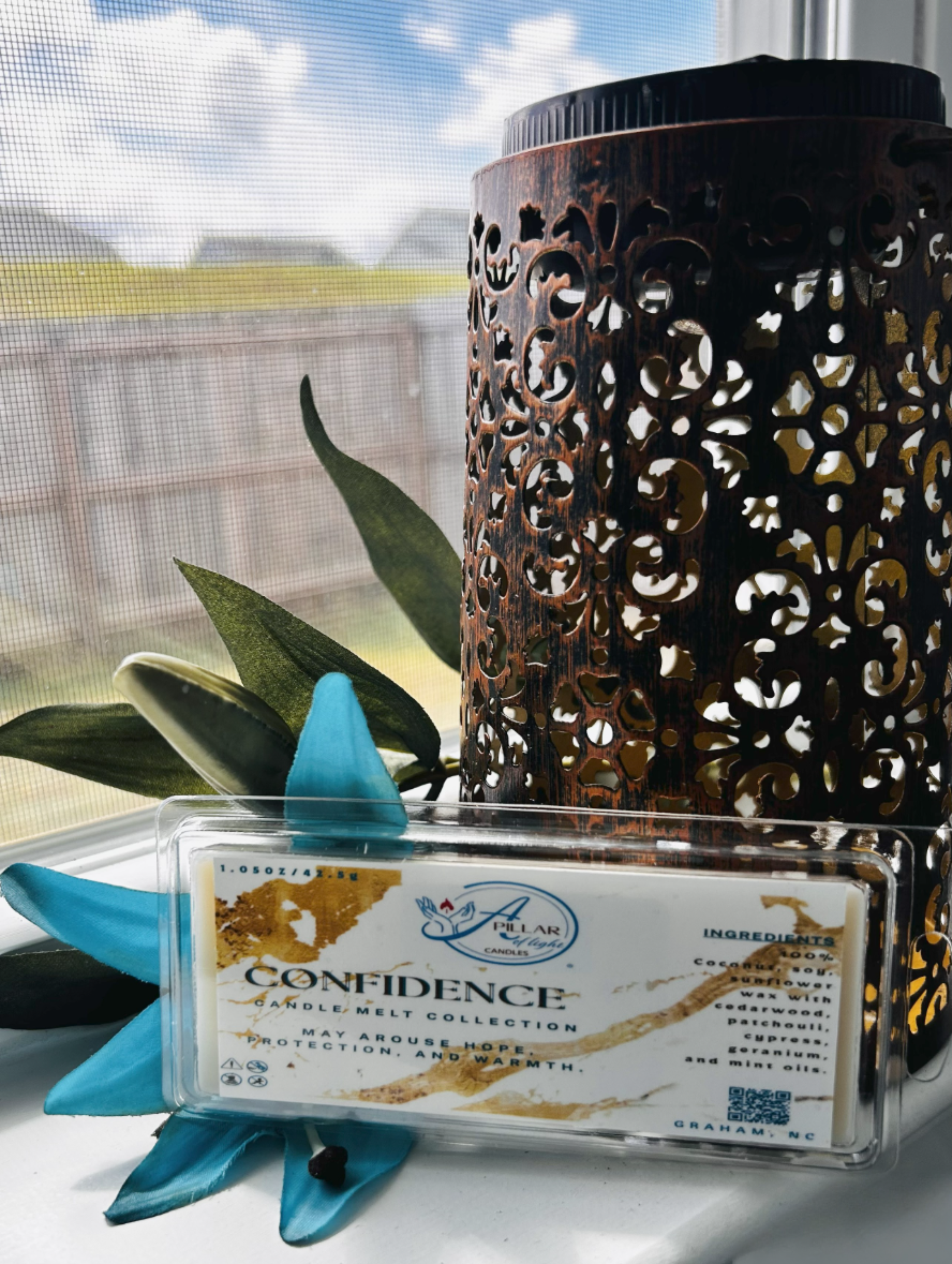 Decorative wax warmer with 'Confidence cedarwood, cypress, patchouli wax melt  on a surface with a window and plant in the background