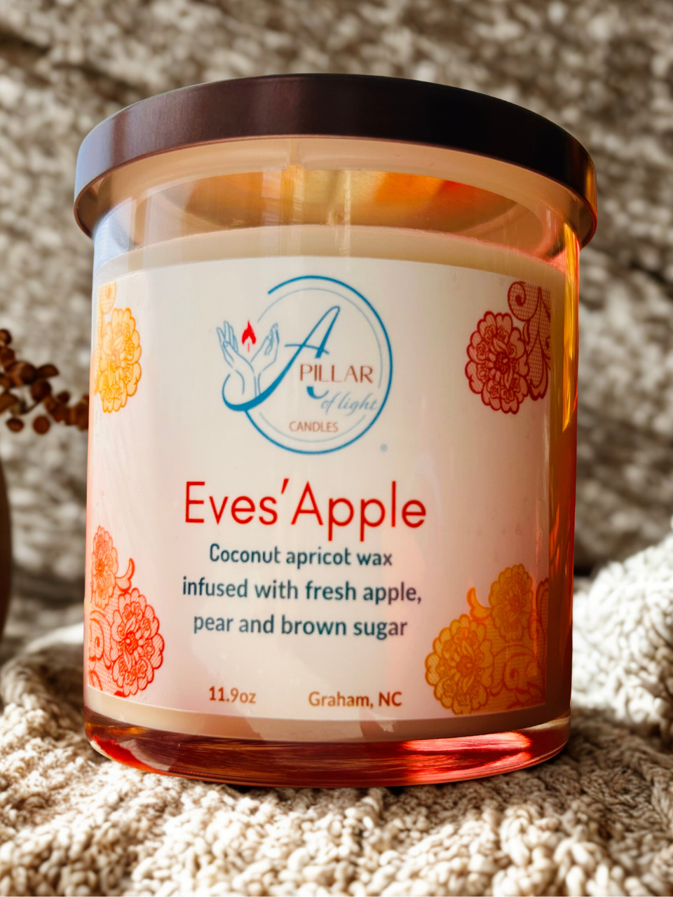 A jar candle labeled 'Eves' Apple' by Pillar of Light Candles, filled with coconut apricot wax and set on a textured beige knit blanket.