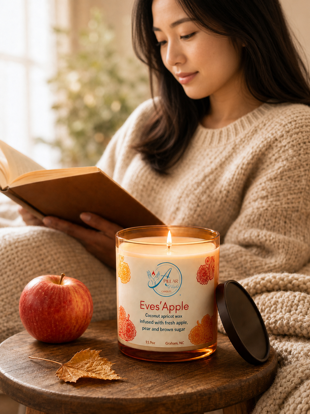 A woman in a beige knit sweater reads a book beside a lit candle labeled 'Eves' Apple' on a wooden table, with a red apple and a dry leaf nearby.