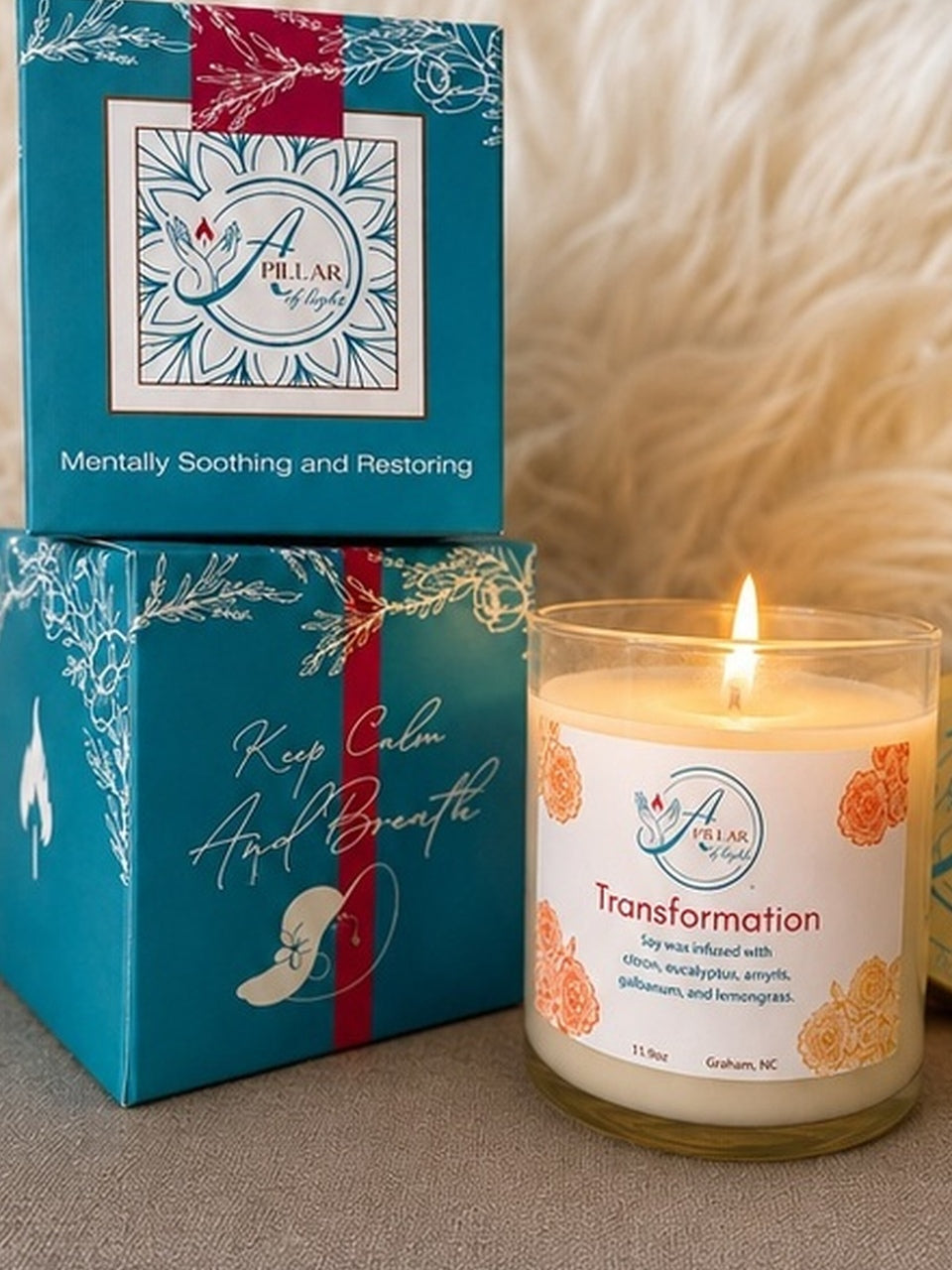 A lit glass jar candle labeled 'Transformation' sits beside stacked teal and white product boxes for Pillar by Aley, with a soft beige fabric background.